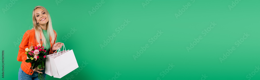 happy and trendy woman with bouquet and shopping bags smiling at camera isolated on green, banner happy and trendy woman with bouquet and shopping bags smiling at camera isolated on green, banner