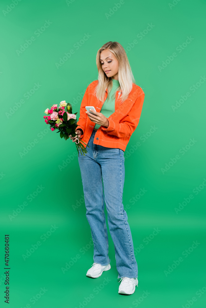 full length view of trendy blonde woman with bouquet messaging on smartphone on green full length view of trendy blonde woman with bouquet messaging on smartphone on green
