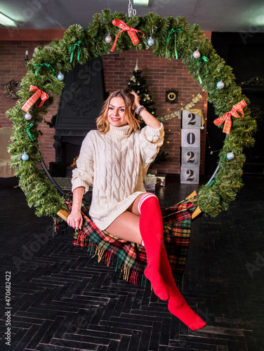 Obraz na płótnie Young pretty woman sitting on a hanging bench in the Christmas decor. The inscription 2022 is behind it. w salonie