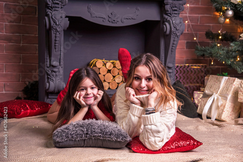 Obraz na płótnie Young pretty mother with daughter lying near the Christmas tree in front of the fireplaceю (New Year, 2022, Christmas concept) w salonie