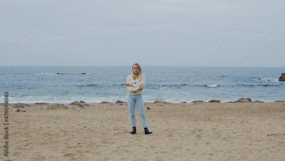 Young blonde woman in casual outfit is standing on rocky ocean coast and looking at camera confidently, beautiful blue tides are on the background, harmony of human and nature, Zoom in, Slow motion. Young blonde woman in casual outfit is standing on rocky ocean coast and looking at camera confidently, beautiful blue tides are on the background, harmony of human and nature, Zoom in, Slow motion.