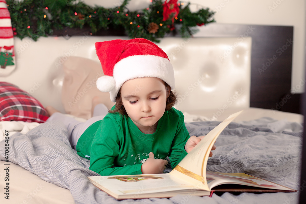 Little cute girl in Santa cap and Christmas pajamas reading a book. Little cute girl in Santa cap and Christmas pajamas reading a book.