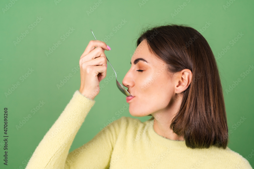 Obraz premium Young woman on a green background in a yellow sweater with appetite eats from a tablespoon, enjoying