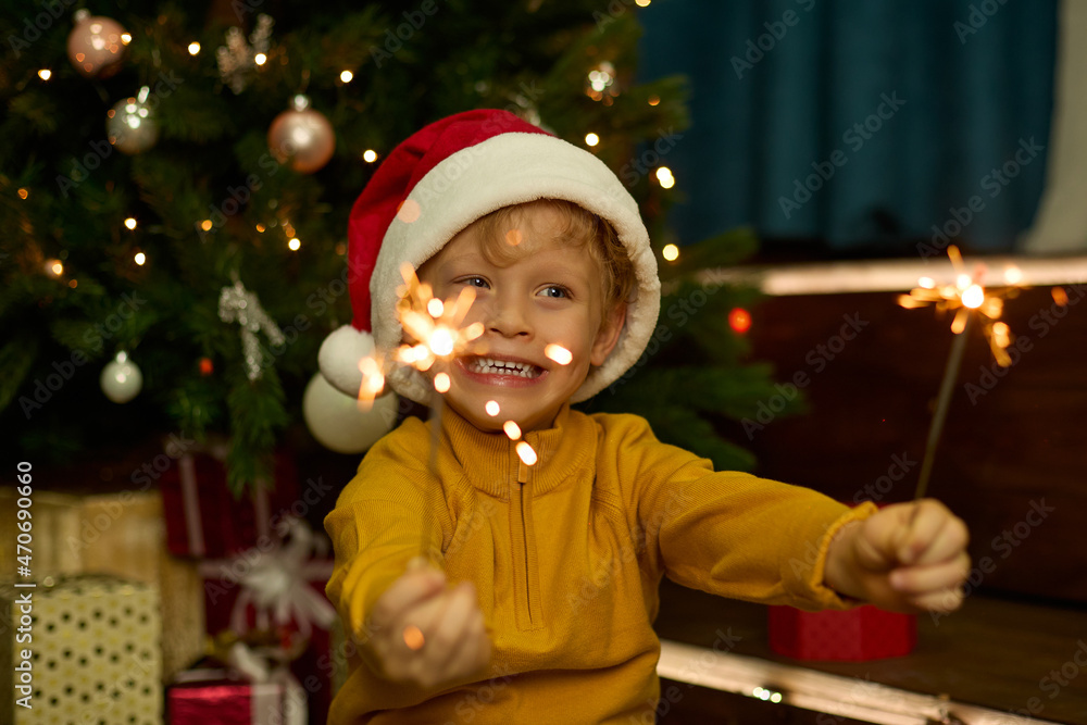 Pretty boy in a New Year is hat, with big blue eyes and a mouth open in surprise holds a sparkling sparkler in his hands against the background of a decorated Christmas tree Pretty boy in a New Year is hat, with big blue eyes and a mouth open in surprise holds a sparkling sparkler in his hands against the background of a decorated Christmas tree