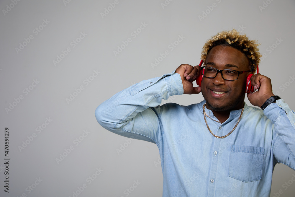 An African-American man is smiling in a blue shirt and glasses, holding red wireless headphones with his hands, listening to music on a white background An African-American man is smiling in a blue shirt and glasses, holding red wireless headphones with his hands, listening to music on a white background