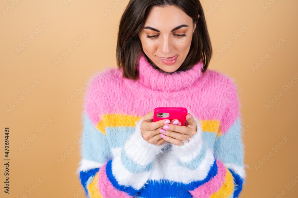 Young woman on a beige background in a knitted sweater with a phone looks at the screen and smiles licks her lip Young woman on a beige background in a knitted sweater with a phone looks at the screen and smiles licks her lip
