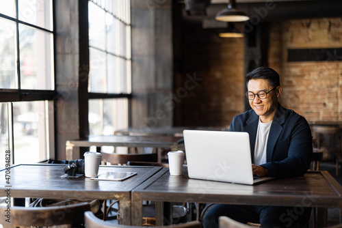Asian businessman typing on laptop during work in cafe. Concept of remote and freelance work. Smiling adult successful man wearing suit and glasses sitting at wooden desk. Sunny day