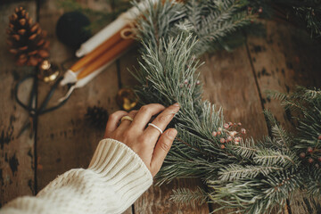 Hand with stylish rings on rustic christmas wreath with pine branches and red berries in snow on rustic old wooden background close up. Atmospheric winter holidays in countryside