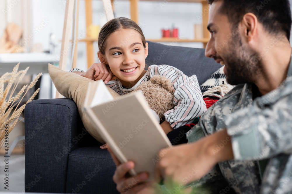 Smiling kid with teddy bear looking at blurred father in military uniform holding book at home Smiling kid with teddy bear looking at blurred father in military uniform holding book at home