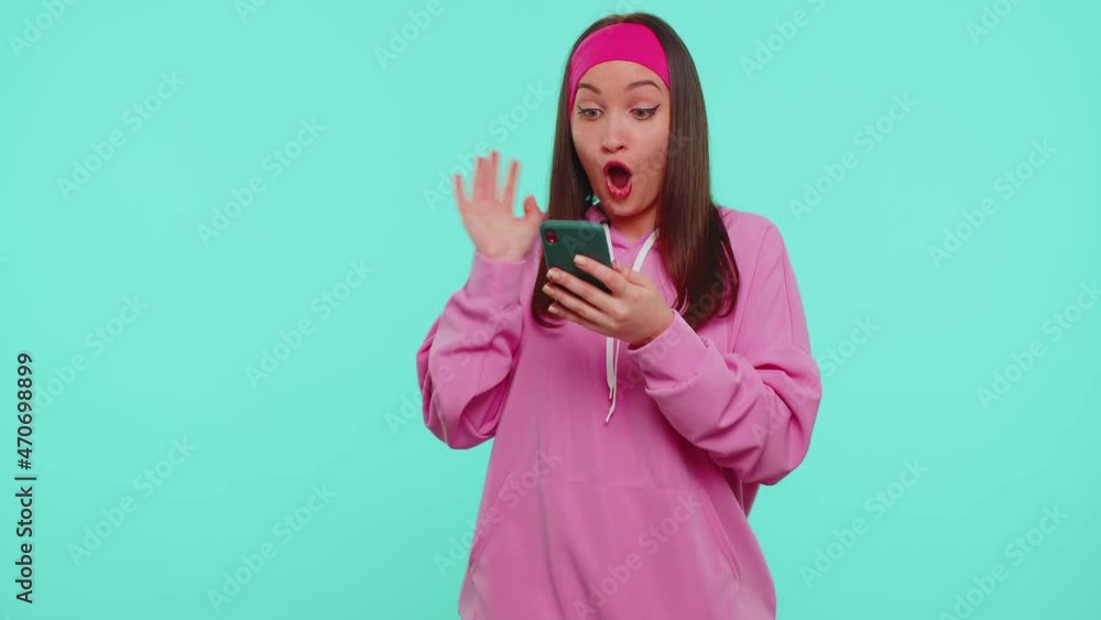 Happy excited joyful stylish teen girl in hoodie use mobile cell phone typing browsing say wow yes found out great big win news doing winner gesture. Young adult woman on blue studio wall background Happy excited joyful stylish teen girl in hoodie use mobile cell phone typing browsing say wow yes found out great big win news doing winner gesture. Young adult woman on blue studio wall background