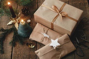 Stylish christmas gifts wrapped in craft paper, candle, scissors, fir branches and bells on rustic wood. Stylish simple scandinavian xmas presents, atmospheric moody image. Eco friendly holidays