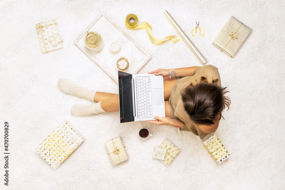 Christmas. Online shopping. Woman in sweater using laptop for making order, buying, sitting on the white carpet among the many wrapped gifts in white gold wrapping paper. Top view. Concept Christmas. Online shopping. Woman in sweater using laptop for making order, buying, sitting on the white carpet among the many wrapped gifts in white gold wrapping paper. Top view. Concept