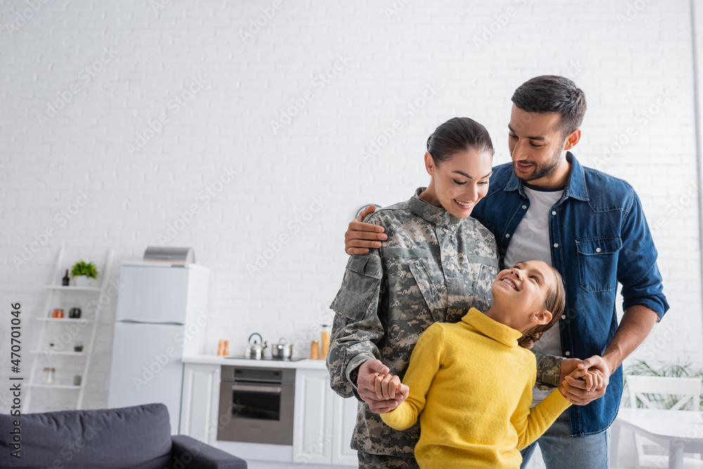 Smiling man hugging wife in military uniform holding hands of daughter at home Smiling man hugging wife in military uniform holding hands of daughter at home