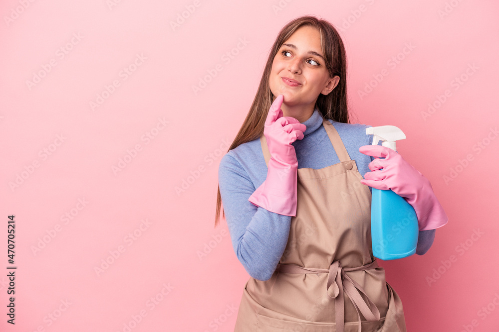 Young caucasian cleaner woman holding spray isolated on pink background looking sideways with doubtful and skeptical expression. Young caucasian cleaner woman holding spray isolated on pink background looking sideways with doubtful and skeptical expression.