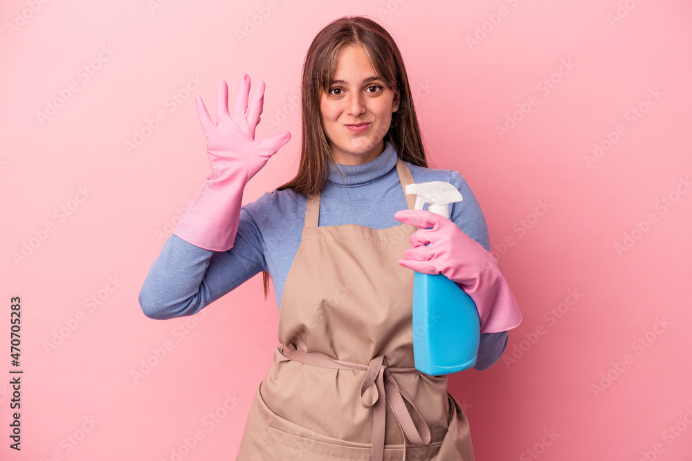 Young caucasian cleaner woman holding spray isolated on pink background smiling cheerful showing number five with fingers. Young caucasian cleaner woman holding spray isolated on pink background smiling cheerful showing number five with fingers.