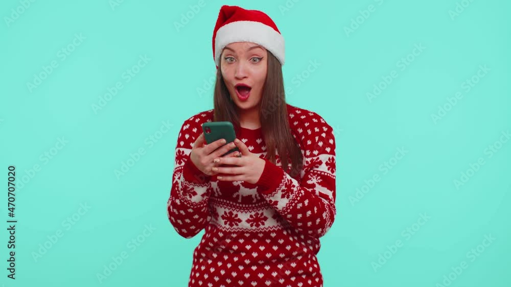 Wow. Girl in stylish Christmas red sweater and Santa hat looking smartphone display sincerely rejoicing win, receiving message with good news, success luck. Woman on blue background. Happy New Year Wow. Girl in stylish Christmas red sweater and Santa hat looking smartphone display sincerely rejoicing win, receiving message with good news, success luck. Woman on blue background. Happy New Year