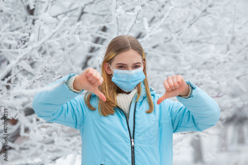 Disappointed young beautiful woman wearing medical mask for protection against coronavirus and gesturing thumb down dislike, standing in a snowy winter park