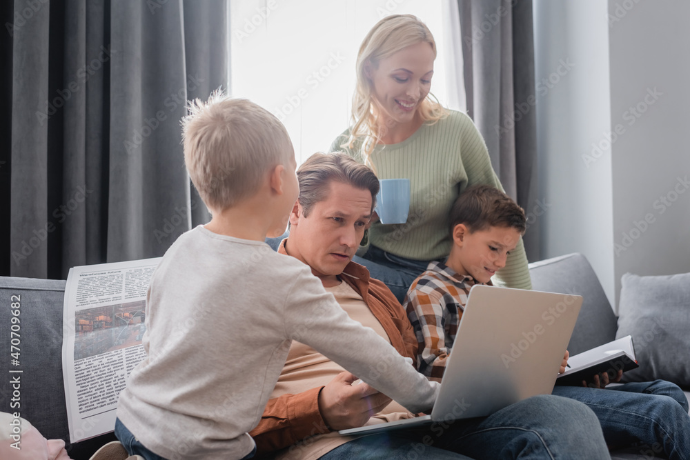 man sitting on couch with laptop near kids with notebook and newspaper and woman standing with cup of tea man sitting on couch with laptop near kids with notebook and newspaper and woman standing with cup of tea