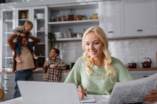 smiling woman with newspaper and pen working near laptop and blurred family in kitchen