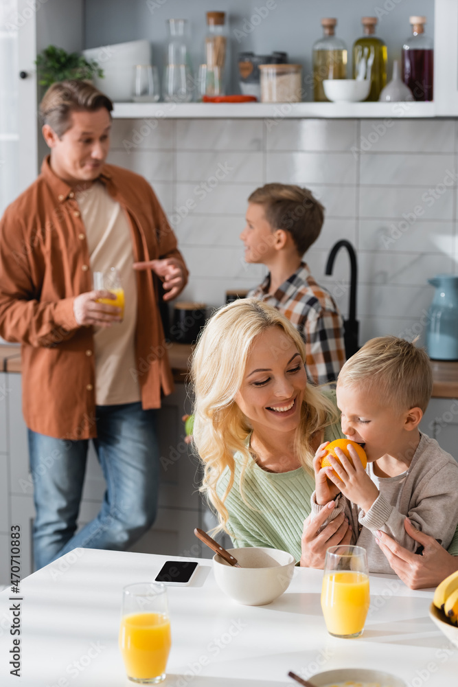pleased kid eating orange near mom and blurred dad with brother in kitchen