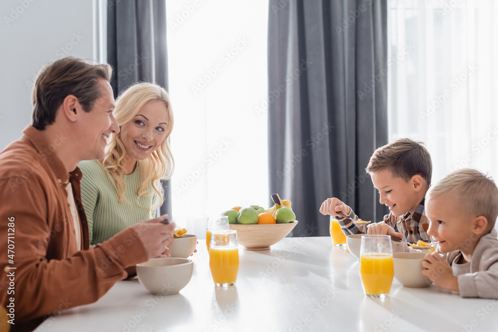 cheerful parents with sons eating corn flakes near orange juice and fresh fruits in kitchen