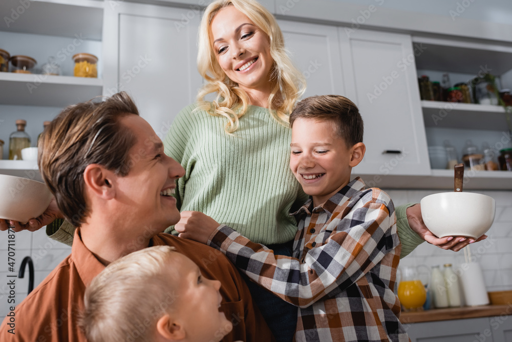 happy woman holding bowls with breakfast near husband and sons in kitchen happy woman holding bowls with breakfast near husband and sons in kitchen