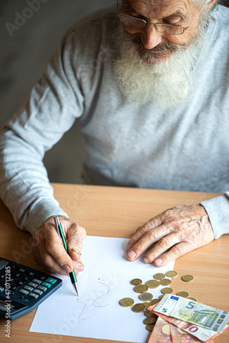Old bearded senior man with calculator and bills counting euro money and writing notes on white sheet of paper.