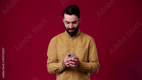 a man writes SMS on the phone to his woman, smiles thoughtfully, he is bearded, black-haired, dressed in a brown shirt, the background is red