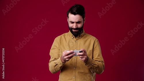 guy plays races on the phone and wins, smiling, joyful, waving his hands, bearded, black-haired, dressed in a brown shirt, background red
