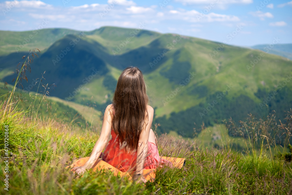 Young happy woman traveler in red dress resting on green grassy hillside on a windy day in summer mountains enjoying view of nature.