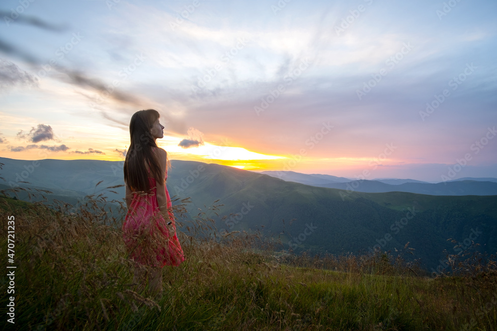 Young woman in red dress walking on hillside meadow on a windy evening in summer mountains