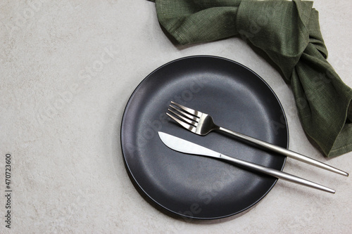Black empty plate with knife and fork with green textile napkin on grey stone background. Table setting. Top view. 