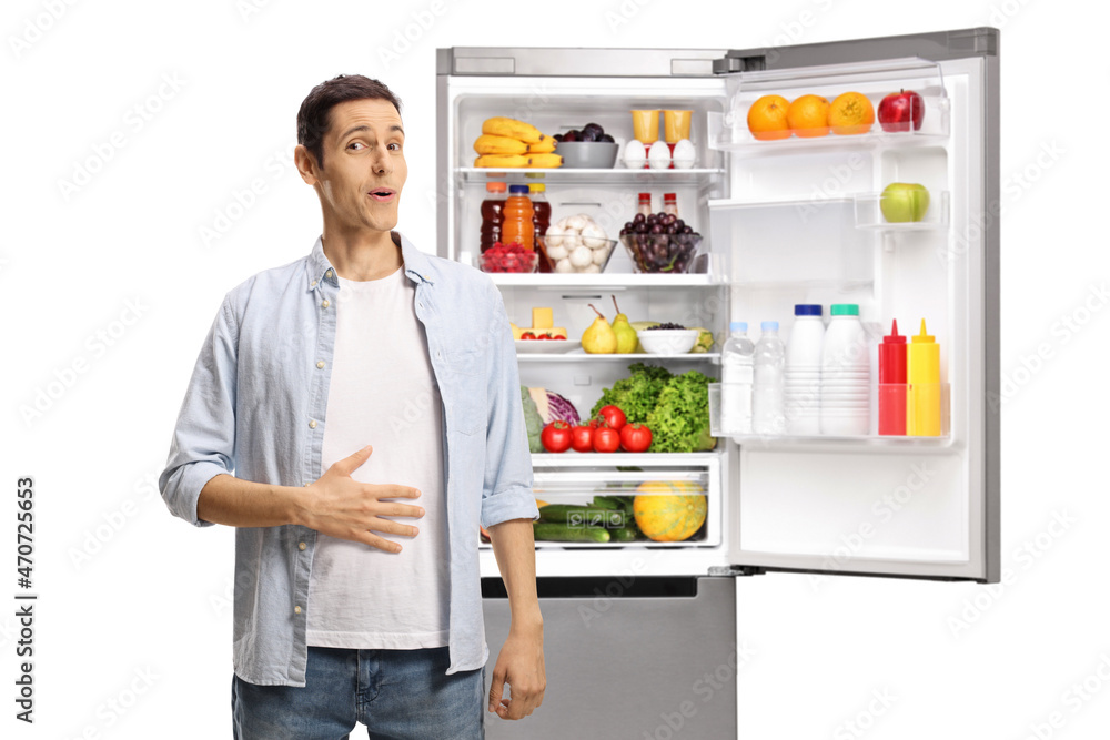 Young man holding his hand on his stomach in front of an open fridge with food Young man holding his hand on his stomach in front of an open fridge with food