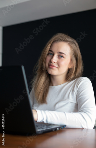 A girl is sitting in front of a laptop, smiling and looking into the lens of the camera at home or in the office. Portrait of a beautiful young girl. Concept of studying or working from home online. 