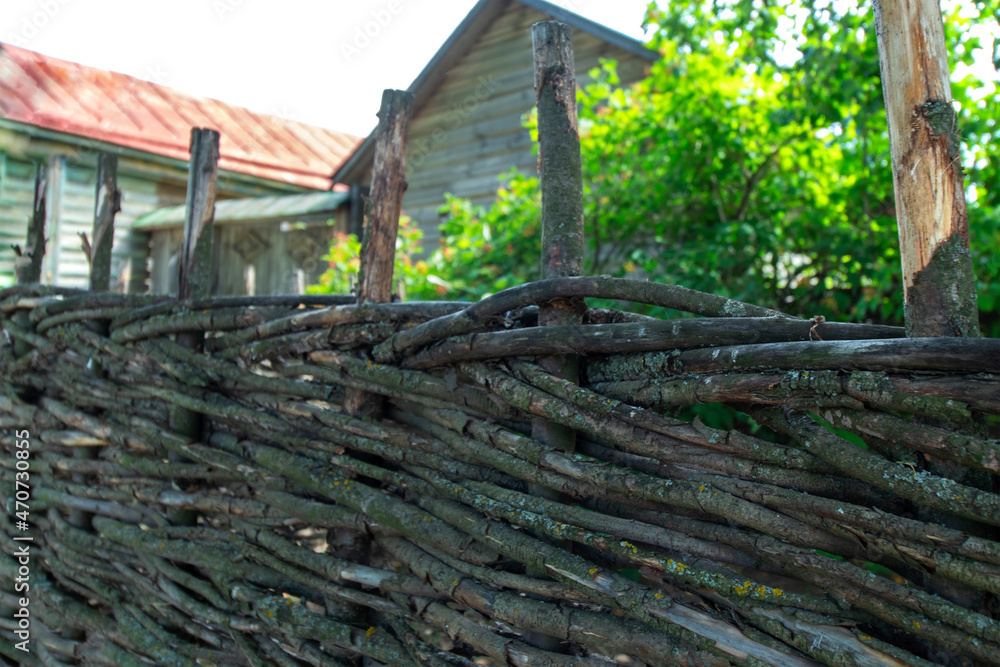 An old wicker fence made of branches against the background of old wooden houses and green bushes. Daylight. Close-up.