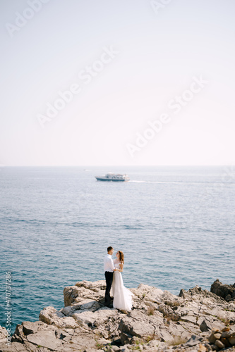 Groom and bride stand on a rocky shore against the background of a ship sailing on the sea