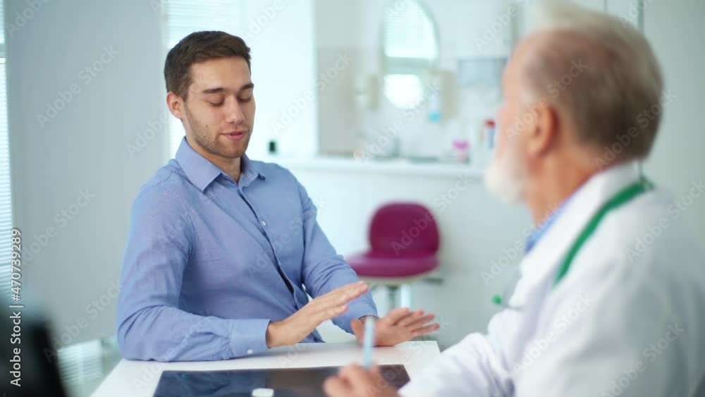 Handsome young man patient talking about problem to mature adult doctor at table in medical office. Focused older senior physician listening to male patient during checkup visit in clinic office. Handsome young man patient talking about problem to mature adult doctor at table in medical office. Focused older senior physician listening to male patient during checkup visit in clinic office.