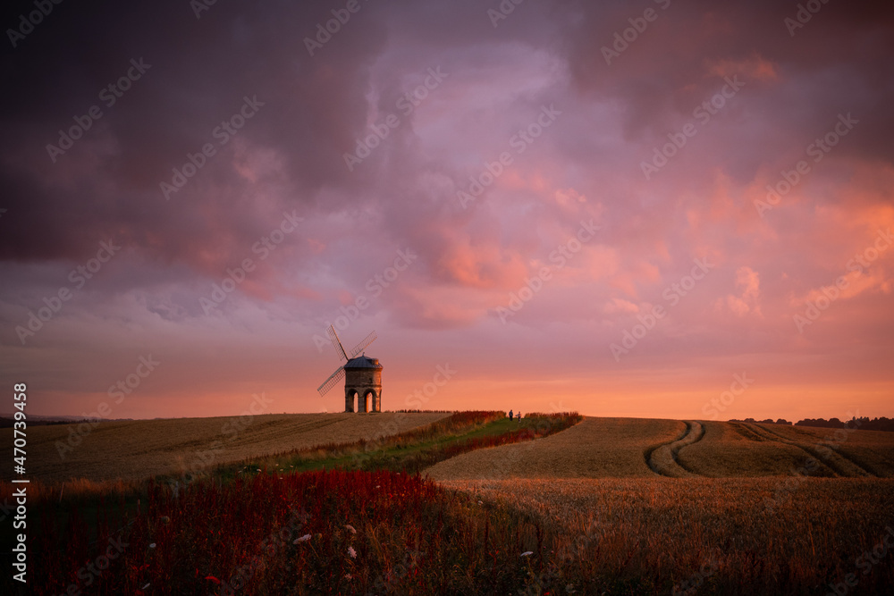 A landmark historic windmill sits on the horizon. Clouds turn pink and dusky purple from the sunset and, in the foreground, a path leads up through the crop fields.