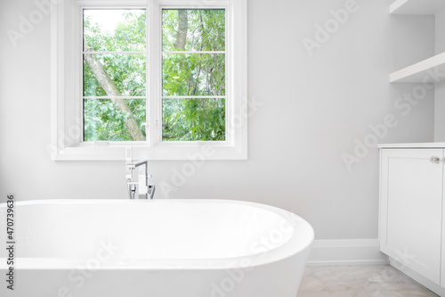 Detail shot of a beautiful white standalone bath tub with chrome hardware and beautiful green trees are seen out of the windows.