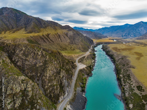 Dark turquoise flow of Katun River and surrounding mountains along the Chuysky Trakt road in the Altai Mountains region of Siberia. Yellow larches and other autumn trees in front on a cloudy day.