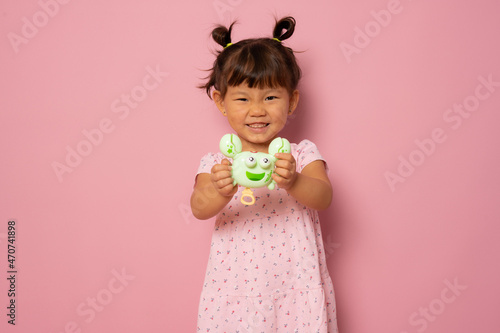 Happy little asian girl in casual dress showing her toy isolated over pink background.