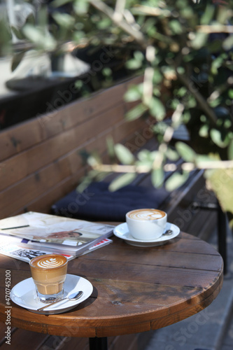 two coffee cups and a magazine are on the table. There is no people in photo. Olives leaves is blur in the photo.