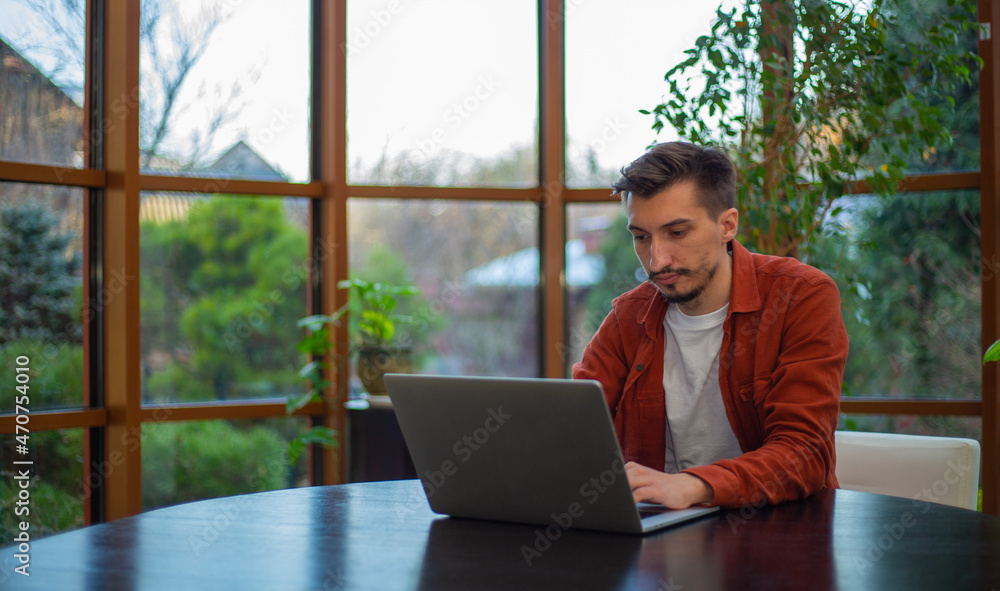 A man in a red shirt works at a laptop at a table during the day against the background of the garden. A man in a red shirt works at a laptop at a table during the day against the background of the garden.