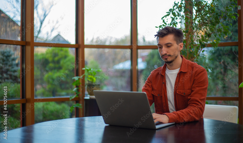 Fototapeta premium A man in a red shirt works at a laptop at a table during the day against the background of the garden.