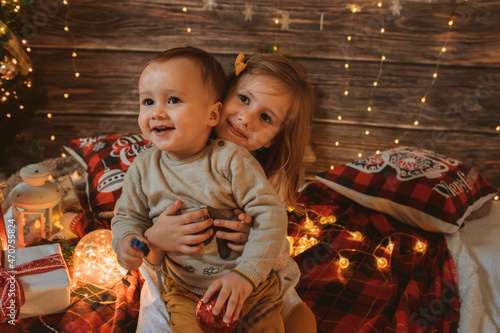 Sister holding her little baby brother in living room decorated by Christmas tree and present gift box, the light give cozy atmosphere. New Year and merry xmas concept