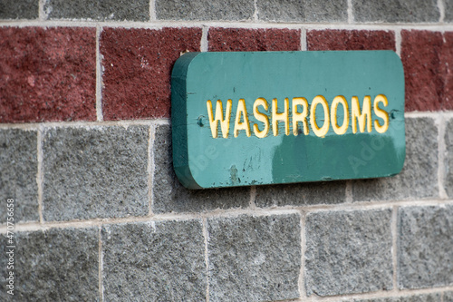 A washroom sign with yellow letters on a green wooden background. The sign is green in color. The wall is grey brick with a red color brick stripe. There's white color mortar between the large bricks.