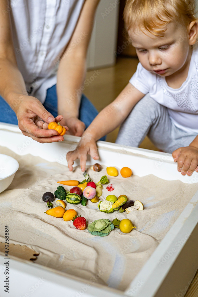 Cheerful baby and mother playing tiny fruit vegetables wooden toys at home kinetic sandbox