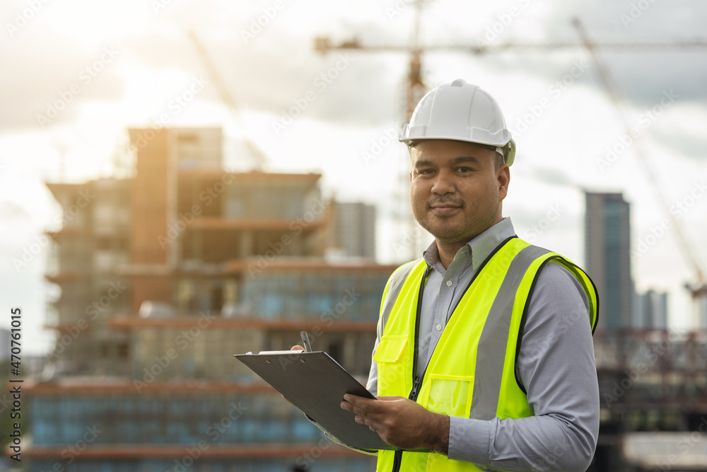 Fototapeta premium Inspector engineer man or architect with clipboard with white safety helmet in city construction site. Checking with checklist on rooftop building construction at capital.