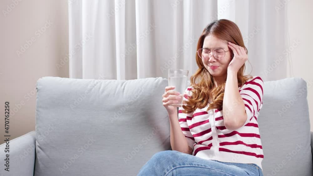 Asian young woman sitting on the sofa drinking water to relax during severe headache. Asian young woman sitting on the sofa drinking water to relax during severe headache.