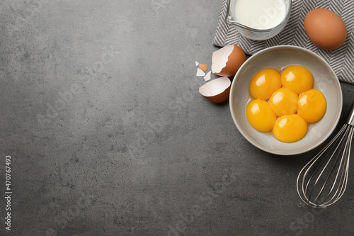 Bowl with raw egg yolks, whisk and jug of milk on grey table, flat lay. Space for text photo